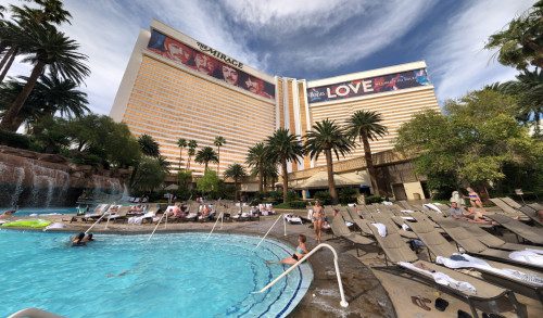 Many visitors relaxing at the pool area with falls surrounded with trees, cabanas, pool-side beds & a casino - hotel building standing at the background named as The Mirage, Las Vegas