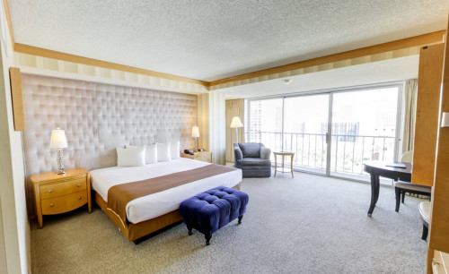 Neatly arranged room of Bally's Las Vegas Hotel and Casino, with cushioned wall panels, cozy bed, single seater couches & a veranda with the city view