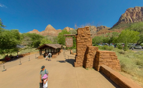 Brick posts & a wide pathway towards the entrance station of Zion National Park in Utah, surrounded by trees & mountains